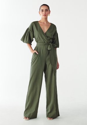 Kombinezon w kolorze oliwkowej zieleni z dekoltem w serek, paskiem w talii, szerokimi nogawkami i luźnymi bufiastymi rękawami. Wykonany z gładkiej tkaniny, posiada boczne kieszenie.
