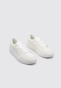 Baskets blanches en cuir lisse avec un bout rond, une semelle plate et des lacets gris clair. Détail de logo subtil sur le côté.
