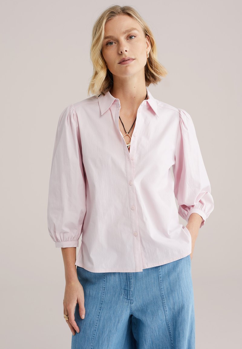 Svetloružová blúzka s nafúknutými rukávmi, zapínaním na gombíky vpredu, golierovým výstrihom a jemnou textúrou, kombinovaná so svetlomodrými širokými nohavicami.