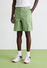Green cargo shorts with a flat front, side pocket, and embroidered logo. Paired with white and gray sneakers featuring red accents.