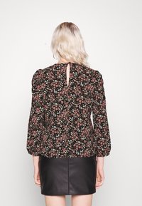 Blouse noire à fleurs avec manches longues bouffantes, ornée de fleurs roses et blanches, détail dos en forme de goutte et silhouette cintrée.