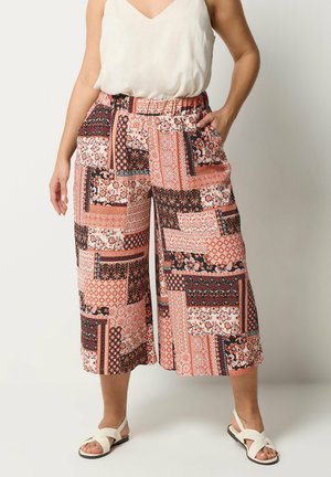 Kobieta nosząca szerokie spodnie sięgające do kostek z pomarańczowym, czarnym i białym kwiatowym i geometrycznym wzorem patchworkowym, w połączeniu z białymi sandałami i bezrękawnikiem.