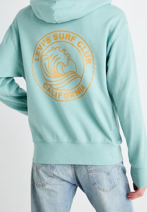 Lichtblauwe hoodie met oranje Levi's Surf Club California-logo met gestileerde golf, gedragen met licht denim jeans, achterkant zichtbaar.