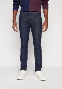 Sötétkék farmernadrág, slim fit fazonnal, öt zsebes kialakítással és kontrasztos narancssárga steppeléssel. Fehér tornacipővel viselve.