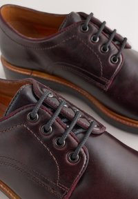 Chaussures en cuir bordeaux foncé à texture lisse, avec des lacets noirs et des coutures détaillées. Elles présentent des œillets ronds pour les lacets et une semelle marron.