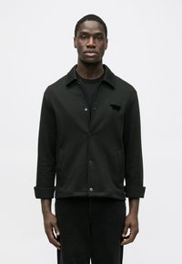 Mustast siledast materjalist nööpidega jakk. Sellel on krae, kaks esist taskut ja peen reljeefne logo rinnal.