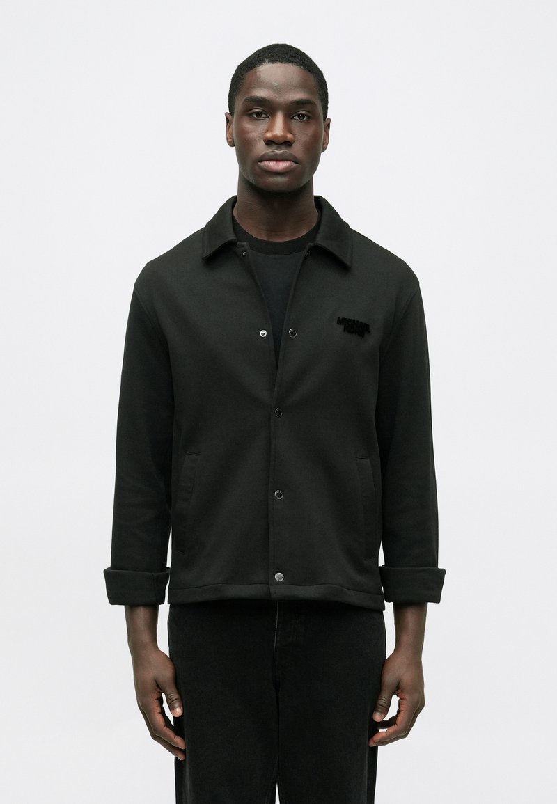 Mustast siledast materjalist nööpidega jakk. Sellel on krae, kaks esist taskut ja peen reljeefne logo rinnal.