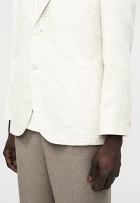 Άνδρας που φοράει κρεμ σακάκι με μοτίβο houndstooth και κουμπιά, συνδυασμένο με μπεζ παντελόνι, στέκεται με το δεξί χέρι χαλαρό στο πλάι.