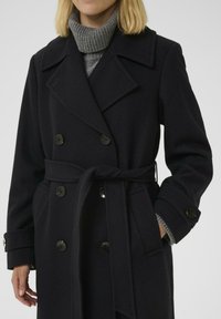 Kvinde iført en sort bæltejakke med dobbeltknapning over en grå strikket turtleneck sweater, hænderne delvist synlige.