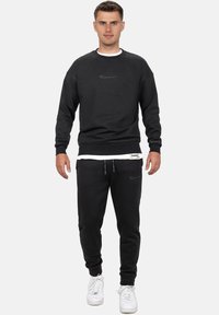 Felpa nera e pantaloni coordinati realizzati in tessuto morbido. La felpa presenta un collo tondo e un ricamo del logo discreto. I pantaloni hanno polsini elastici.