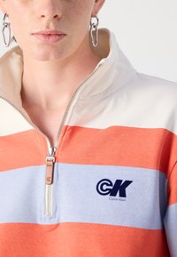 Calvin Klein kvart-zip genser med oransje og lys blå horisontale striper, med en sølvglitrende glidelås og logo pute på brystet.
