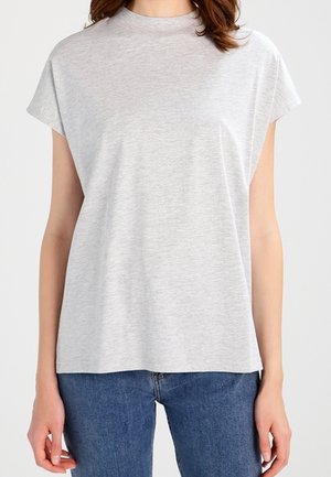 Kobieta w prostym jasnoszarym topie z krótkim rękawem, luźno włożonym do niebieskich dżinsów, stojąca na jednolitym tle.