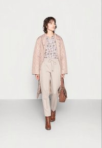 Casaco longo acolchoado bege sobre uma blusa floral, combinado com calças bejes claras e botas de tornozelo castanhas. Bolsa castanha como acessório.