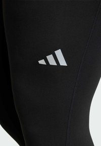 Black activewear leggings with a smooth texture, featuring a white three-stripe logo on the upper thigh and visible flatlock stitching.