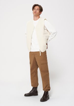 Beżowa bezrękawnik na białej bluzie, zestawiona z brązowymi spodniami cargo i czarnymi sznurowanymi butami; minimalistyczny, funkcjonalny design.