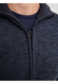 Dunkelblaues Reißverschluss-Sweatshirt mit strukturiertem Strickmuster, geripptem Kragen und sichtbarem Reißverschluss. Über einem einfarbigen dunkelblauen Shirt getragen.