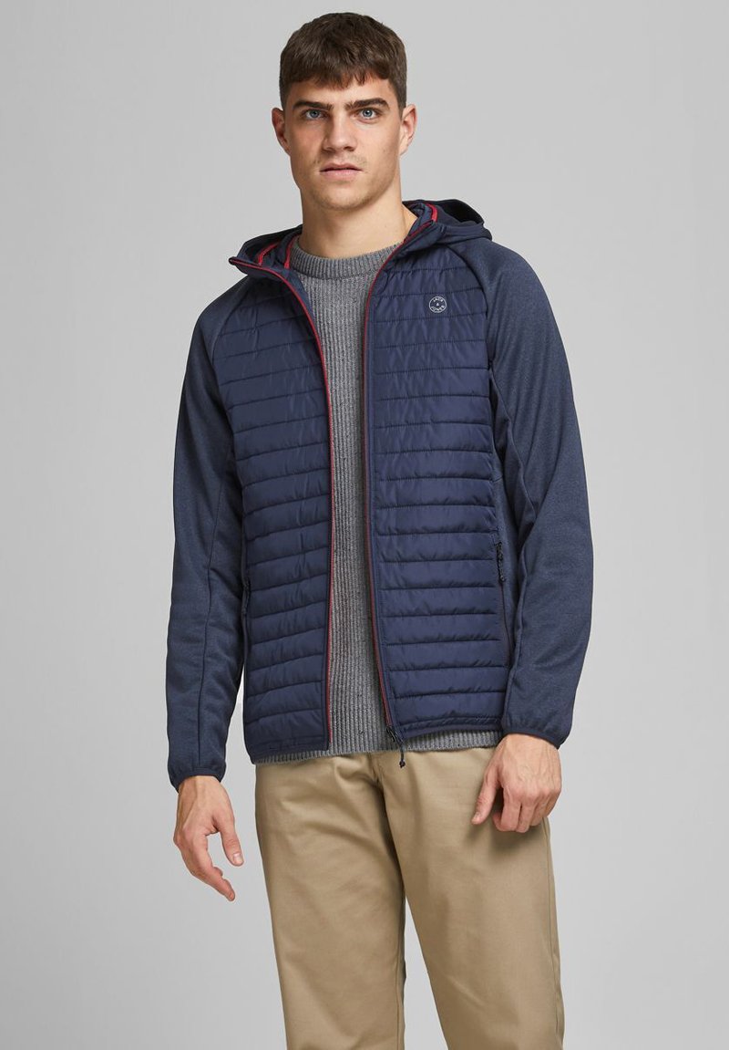Navy puffer jacket with textured sleeves, red zipper accents, and a fitted hood, paired with a gray sweater and beige pants.