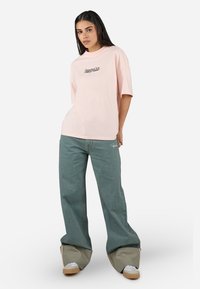 Rosa oversized t-skjorte med brodert tekst, kombinert med løse, grønne denimbukser med oppbrett og hvite sneakers med beige accenter.