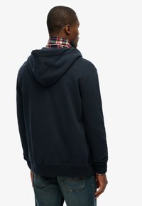 Sweat à capuche bleu marine avec un ourlet et des poignets côtelés, superposé à une chemise à carreaux rouge. Texture lisse et coupe décontractée.