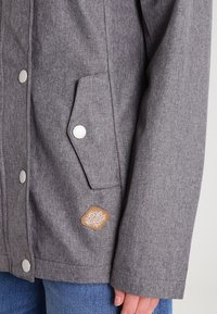 Gray jacket with textured fabric, snap buttons, a side pocket, and a brown logo patch. Features a clean, utilitarian design.