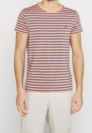 Homme portant un t-shirt à manches courtes à rayures horizontales orange et bleu et un pantalon de couleur claire, debout devant un fond uni.