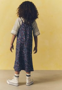 Dunkelblaues Kleid mit floralem Muster und dünnen Trägern, darüber ein graues T-Shirt. Kombiniert mit weißen High-Top-Sneakern und gestreiften Socken.