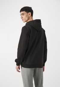 Zwarte hoodie met een ontspannen pasvorm, ribgebreide manchetten en zoom, gedragen met grijze sweatpants. De stof lijkt zacht en glad.