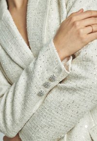 Blazer bianco texturizzato con paillettes scintillanti e tre bottoni decorativi argentati sul polsino. Tessuto morbido con un design su misura.