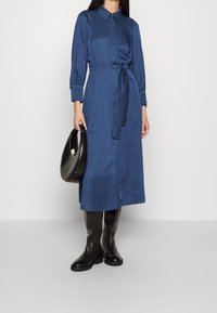 Lång blå skjortklänning med ett strukturerat mönster, krage, midjebälte och knäppta ärmar, i kombination med knähöga svarta stövlar och en svart väska.