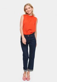 Blusa rossa senza maniche con scollatura arricciata, jeans dritti blu scuro con risvolti e sandali rosa con cinghie. Outfit semplice e casual.