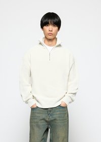 Light cream pullover made from textured fabric with a half zip, featuring ribbed cuffs and hem, worn over a white t-shirt and blue jeans.