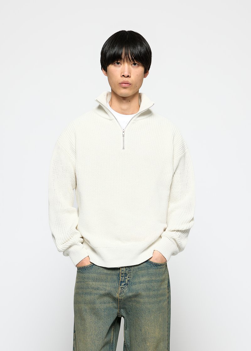 Light cream pullover made from textured fabric with a half zip, featuring ribbed cuffs and hem, worn over a white t-shirt and blue jeans.