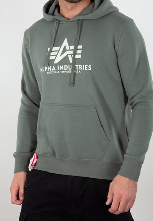 Mężczyzna w oliwkowozielonej bluzie z kapturem Alpha Industries z przednią kieszenią oraz białym napisem „Knoxville, Tennessee U.S.A.” na piersi.