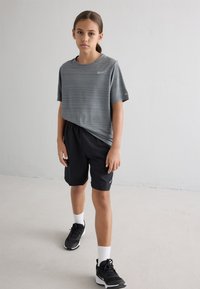 Grau gestreiftes Nike T-Shirt mit kurzen Ärmeln, kombiniert mit schwarzen Shorts. Schwarze Sportschuhe und weiße Socken runden das Outfit ab. Einfarbiger Hintergrund.