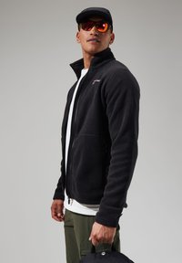 Black fleece jacket with zip, high collar, and front pockets. Worn over a white shirt. Model carries a black bag. Sunglasses add reflective detail.