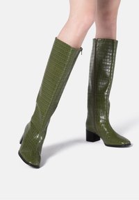 Bottes hautes vertes avec une texture brillante embossée effet croco, bout carré et talon bloc. Fermeture éclair latérale pour un enfilage facile.