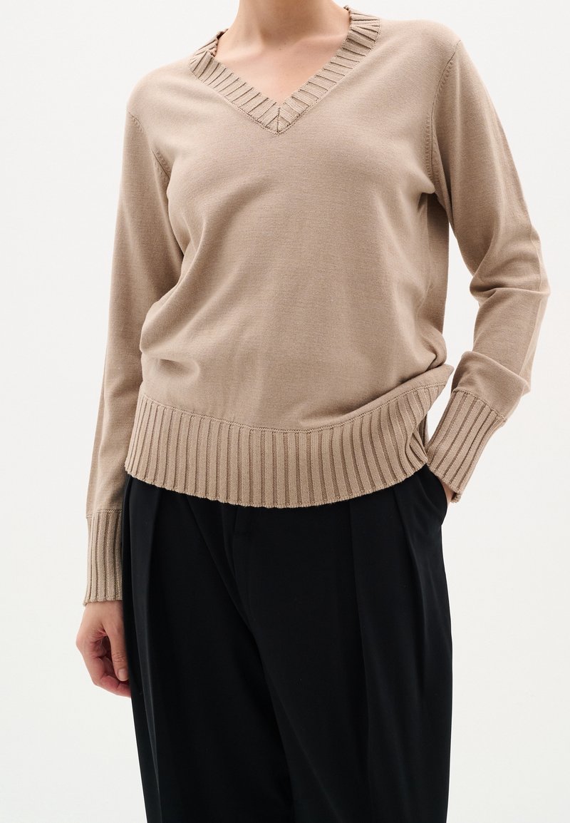 Beige V-ringad tröja tillverkad av mjukt tyg, med en texturerad ribbad halsringning och fåll, långa ärmar och en avslappnad passform.