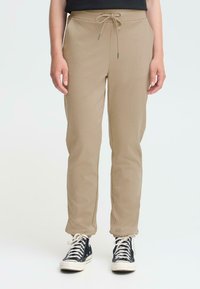 Beige joggingsbroeken van zacht materiaal, met een elastische tailleband en een trekkoord, taperend bij de enkels. Draag ze met zwarte sneakers.