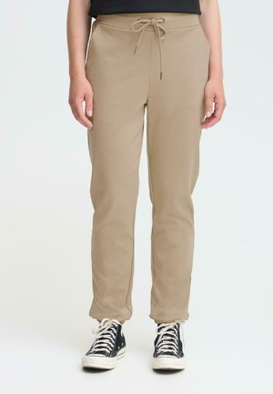 Beige jogger-housut, jotka on valmistettu pehmeästä kankaasta ja joissa on elastinen vyötärö nauhalla, kapenevat nilkoissa. Yhdistetty mustiin lenkkareihin.