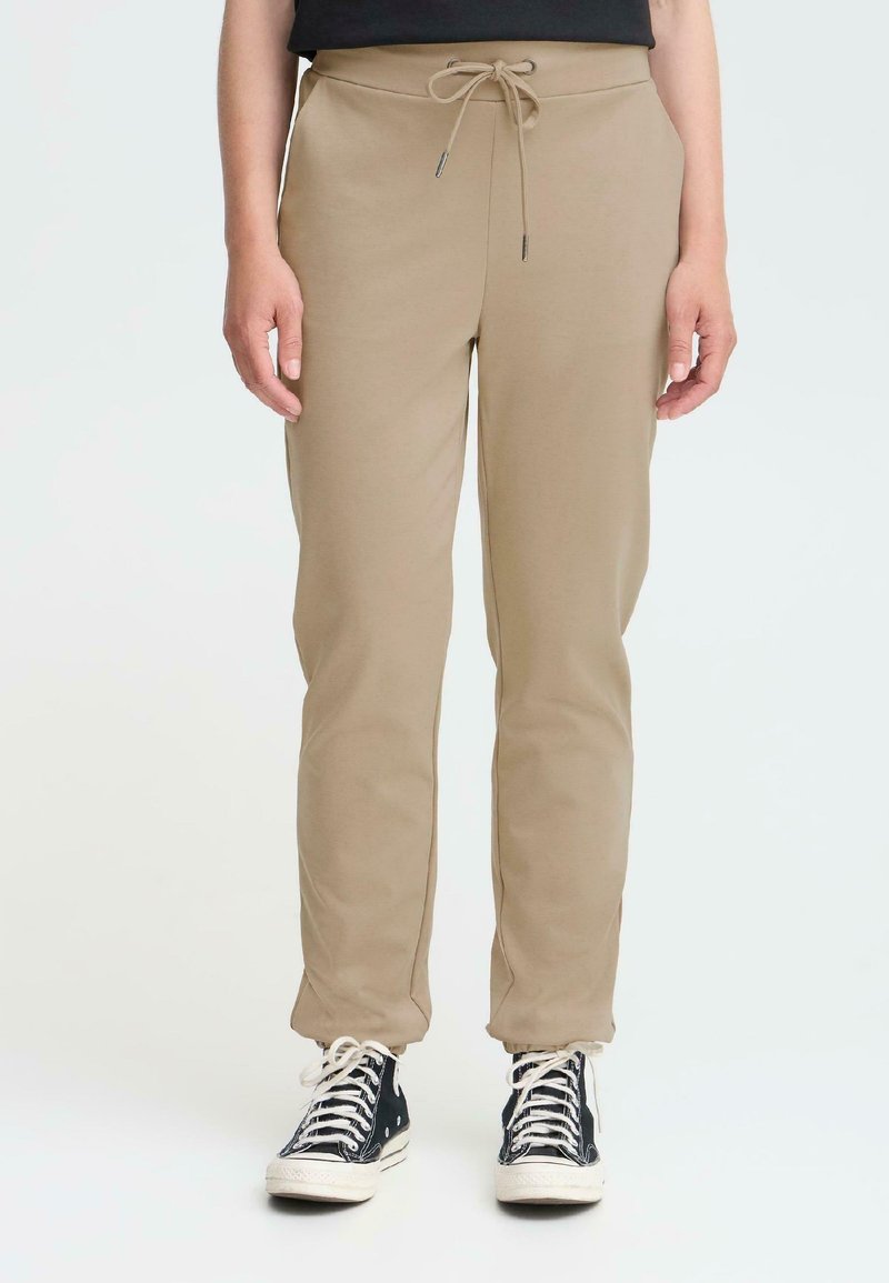 Beige joggingsbroeken van zacht materiaal, met een elastische tailleband en een trekkoord, taperend bij de enkels. Draag ze met zwarte sneakers.