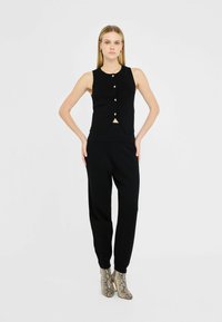 Black sleeveless top with front buttons and a slight V-shape, paired with matching black pants. Model wears gray snake-skin ankle boots.