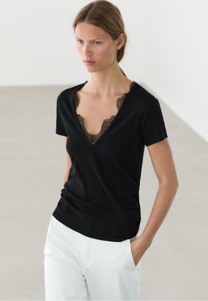 Kvinde iført en sort T-shirt med V-hals med sort blonde detaljer og hvide bukser, stående med den ene hånd i lommen i et ensfarvet lyst rum.