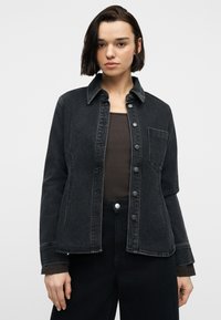 Νεαρή γυναίκα με κοντά σκούρα μαλλιά, φορώντας μαύρο τζιν μπουφάν πάνω από σκούρα μπλούζα και μαύρο παντελόνι, στέκεται μπροστά σε απλό φωτεινό φόντο.