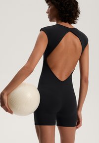 Zwarte mouwloze bodysuit met een diepe V-rug, gemaakt van een glad materiaal. Het model houdt een gestructureerde, lichtgekleurde bal met golvende patronen vast.