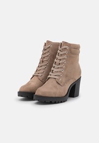 Botas de ante marrón hasta el tobillo con un tacón negro grueso, con ojetes de metal y cordones, y un cuello ligeramente acolchado para mayor comodidad.
