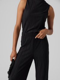 Combinaison noire plissée avec une coupe sans manches, un col montant et une taille élastique. Le modèle tient un sac noir texturé.