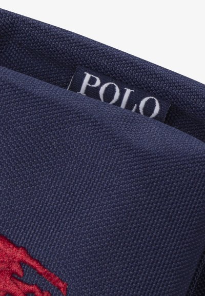 Artėjantis vaizdas tamsiai mėlynos spalvos audinio su baltu „POLO“ etiketės užrašu ir raudonu siuvinėtu arkliu su raituojančiu žmogumi logotipu.
