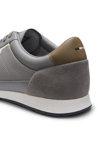 Chaussure de sport grise avec panneau latéral texturé, talon en daim, col en cuir marron et semelle blanche avec adhérence noire, vue par l'arrière.