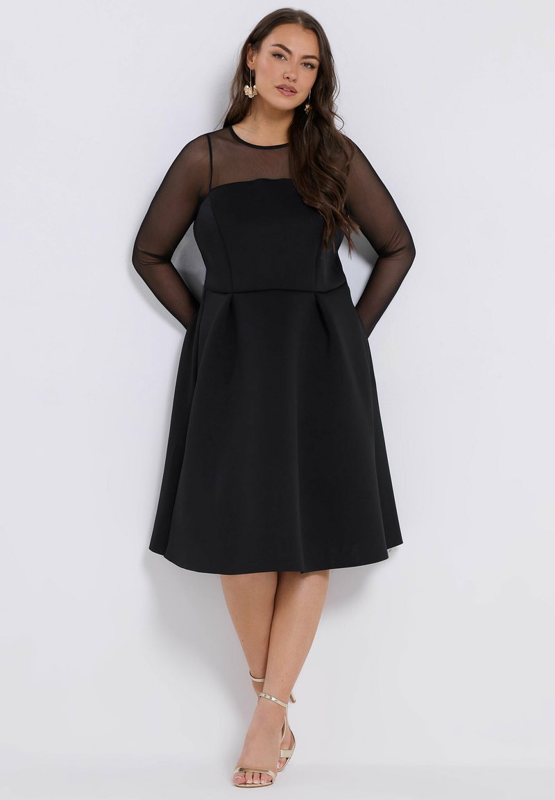 Nainen mustassa polvijalan mekossa, jossa on läpinäkyvät pitkät hihat, seisoo kädet takana, jalassa kultaiset remmikengät ja korvakorut.