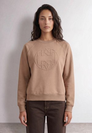 Femme aux longs cheveux foncés portant un sweat-shirt beige avec un logo circulaire embossé et un pantalon marron foncé, debout contre un mur uni.